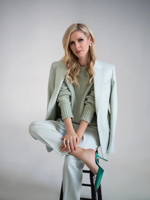 Blonde woman in light green suit poses seated on a stool in a studio setting, showcasing a modern and elegant fashion aesthetic.