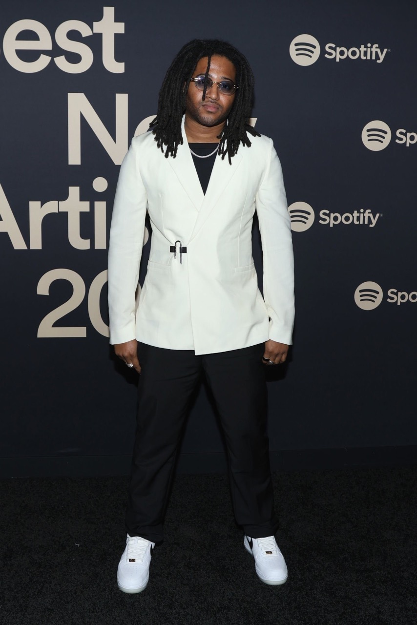 Man with dreadlocks poses in white suit and white sneakers on red carpet at an event with Spotify logo backdrop.