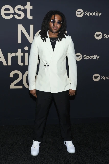 Man with dreadlocks poses in white suit and white sneakers on red carpet at an event with Spotify logo backdrop.