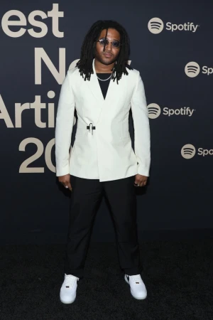 Man with dreadlocks poses in white suit and white sneakers on red carpet at an event with Spotify logo backdrop.