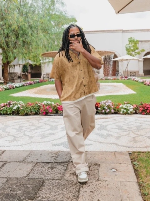 Man in a patterned shirt and khaki pants walks on a stone path in a formal garden with a fountain, flowers, and whitewashed buildings.