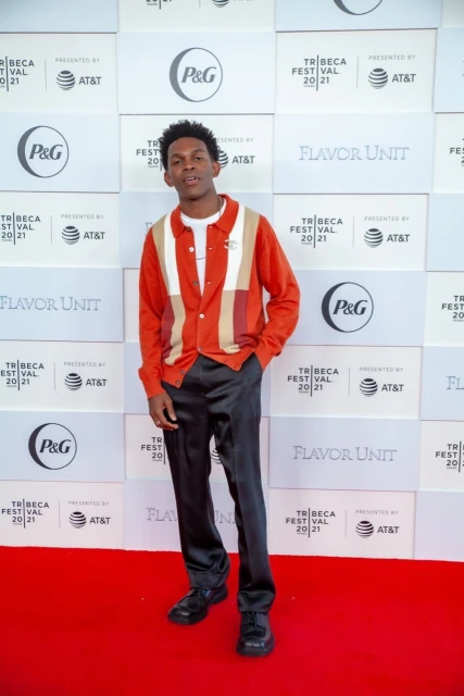 A young man in a patterned shirt and black pants stands on a red carpet against a branded backdrop at the Tribeca Film Festival.