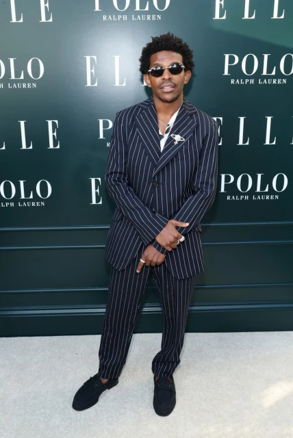 Adult male in pinstripe suit and sunglasses poses in front of a backdrop with fashion brand logos. Daytime, indoor setting.