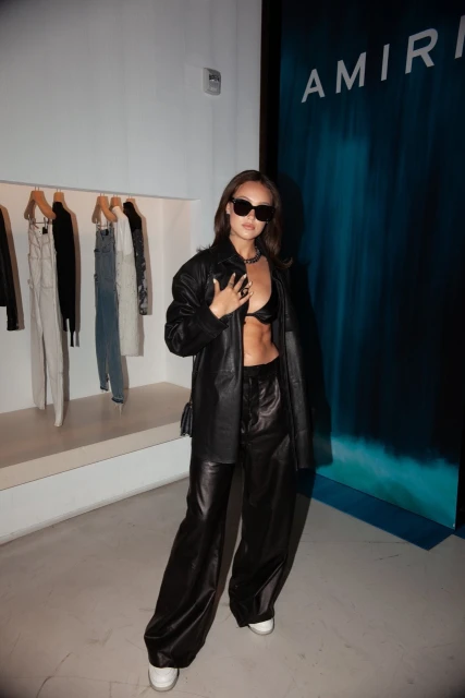 Young woman in a black leather jacket and pants posing indoors next to clothing rack and a blue branded background, wearing sunglasses.