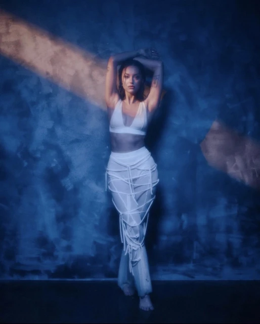 A woman in a white two-piece outfit poses against a blue textured wall with hands above her head in a studio setting.