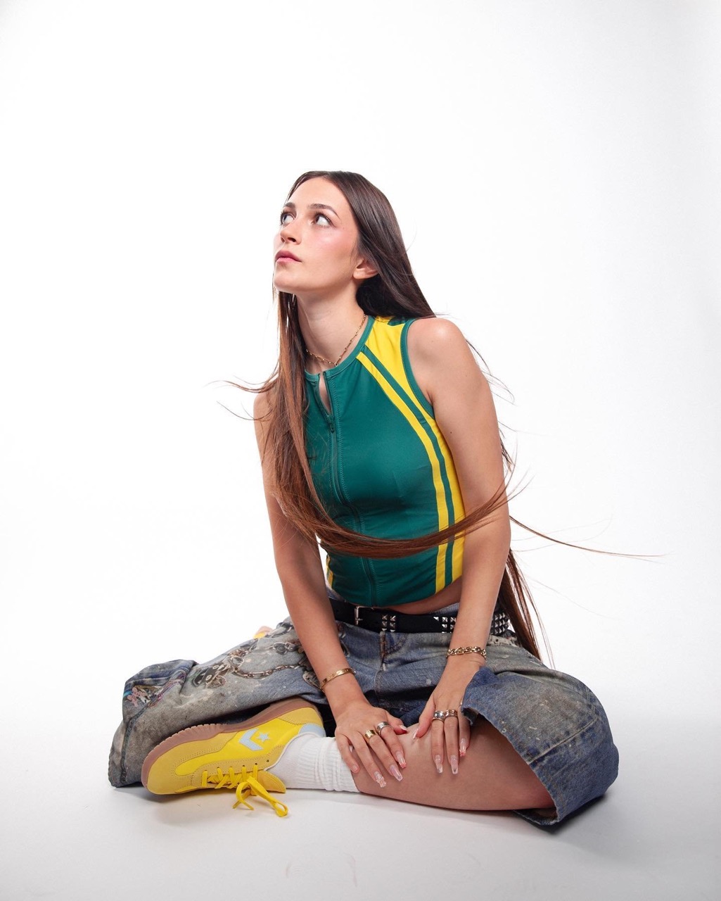 A young woman with long brown hair sits cross-legged looking up in a green and yellow tank top, jean skirt, and yellow sneakers in a white studio.