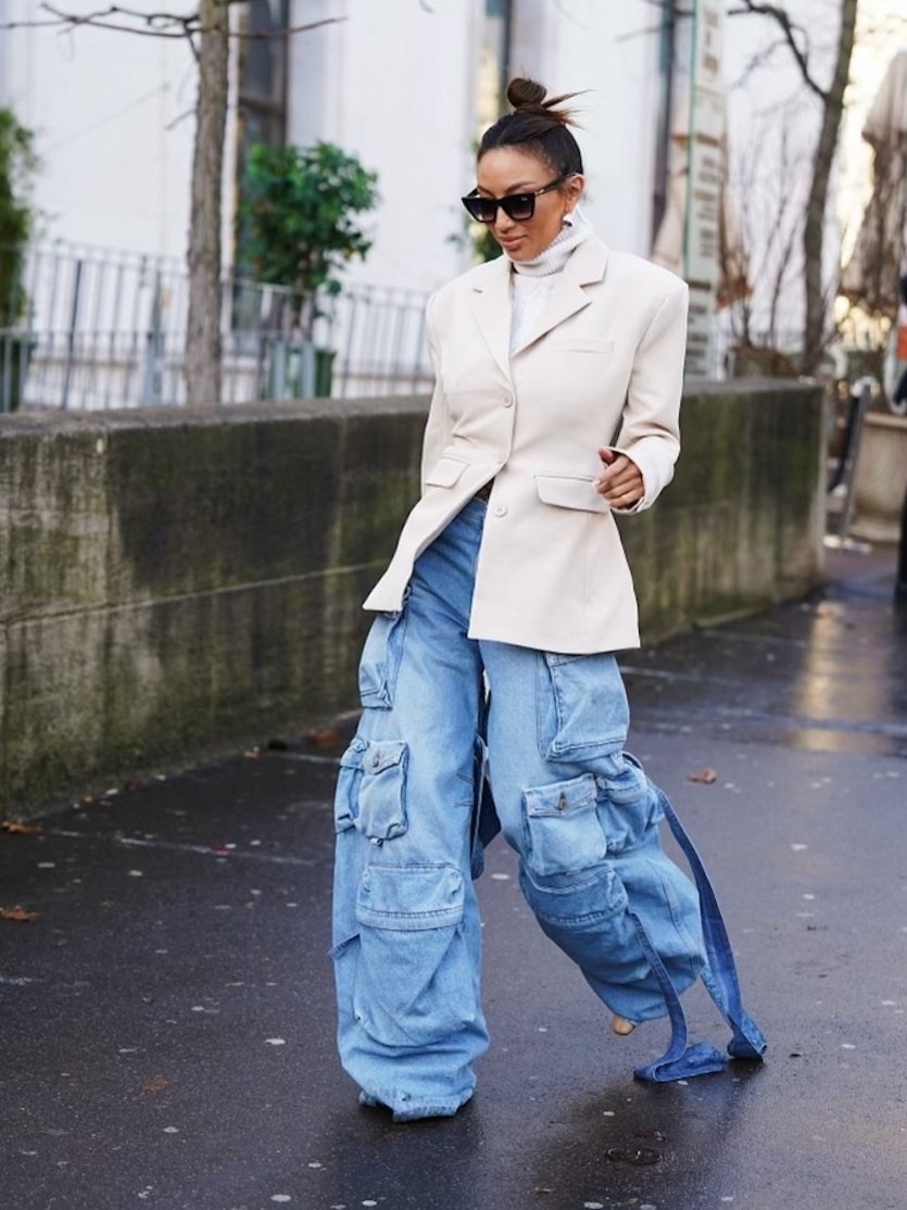 Fashionable adult woman wearing oversized light blue denim cargo pants and beige blazer poses in a city street with trees in the background.