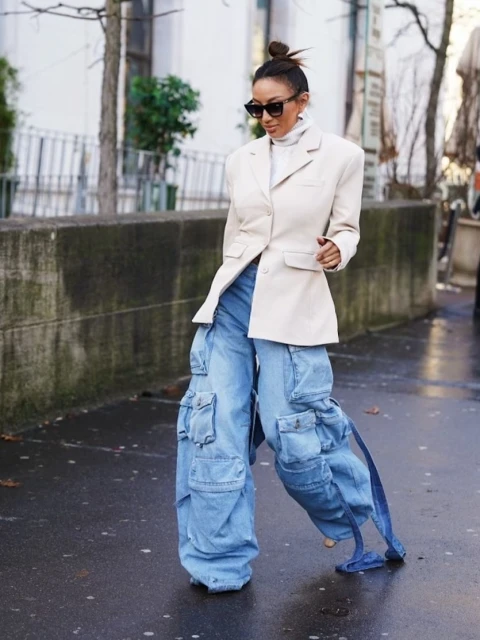 Fashionable adult woman wearing oversized light blue denim cargo pants and beige blazer poses in a city street with trees in the background.