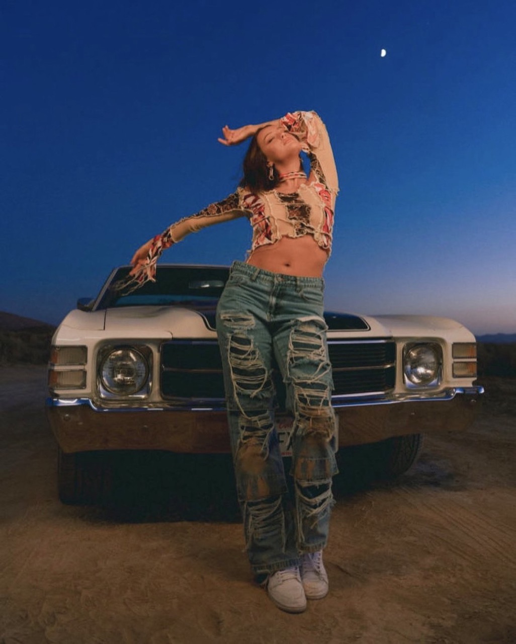 Woman in ripped jeans poses confidently in front of a vintage car against a twilight desert backdrop with a visible moon.