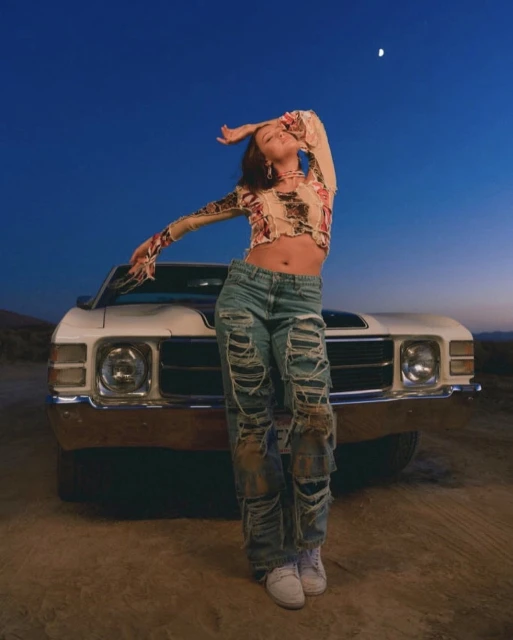 Woman in ripped jeans poses confidently in front of a vintage car against a twilight desert backdrop with a visible moon.