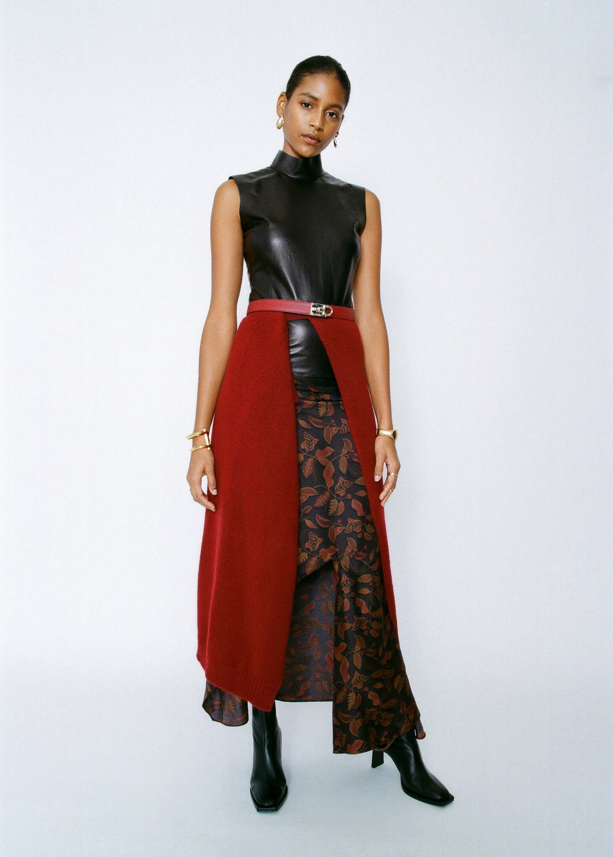 Fashionable woman wearing a black leather top, red vest, and patterned skirt, posing in a studio with a white background.