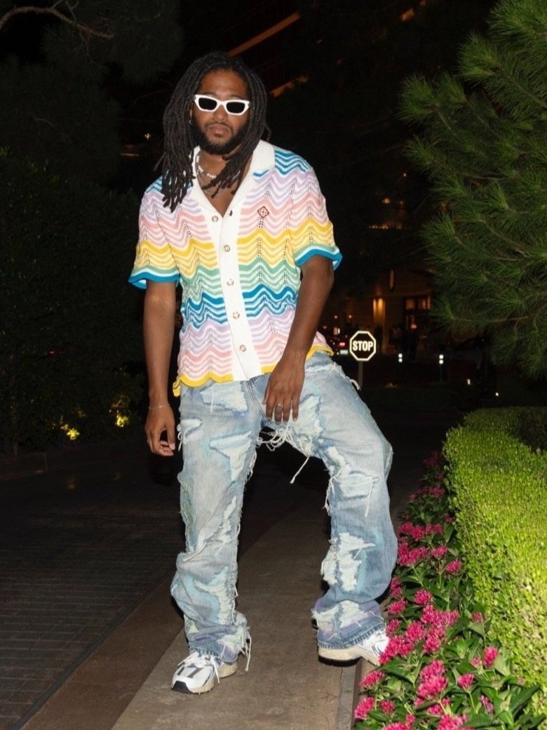 Adult male wearing sunglasses and a colorful shirt with ripped jeans stands in a nighttime urban setting near greenery.
