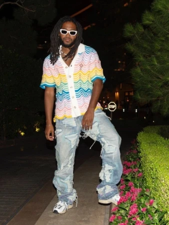 Adult male wearing sunglasses and a colorful shirt with ripped jeans stands in a nighttime urban setting near greenery.