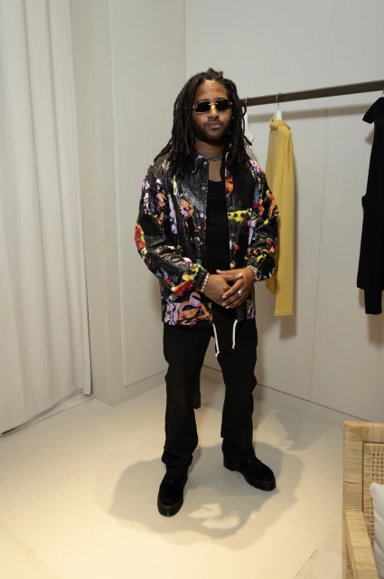 A stylish adult male with dreadlocks and sunglasses poses indoors wearing a floral-print jacket, black pants, and black boots.