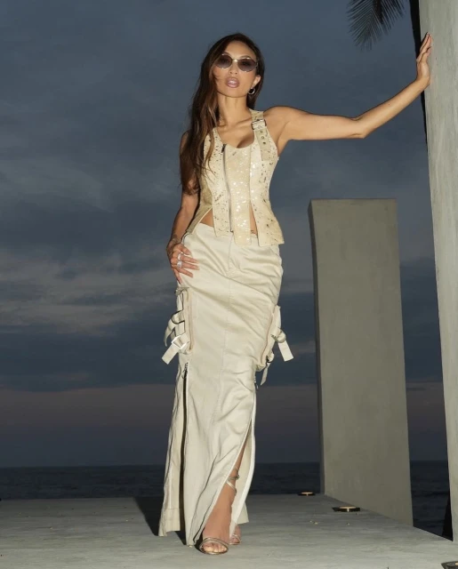 Woman poses in cream corset top and long skirt with palm tree and dusk sky background, showcasing fashion and outdoor elegance.