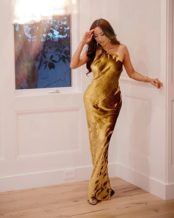 Woman in a long gold metallic one-shoulder silk dress posing indoors near a white wall and window styled by Marissa Pelly
