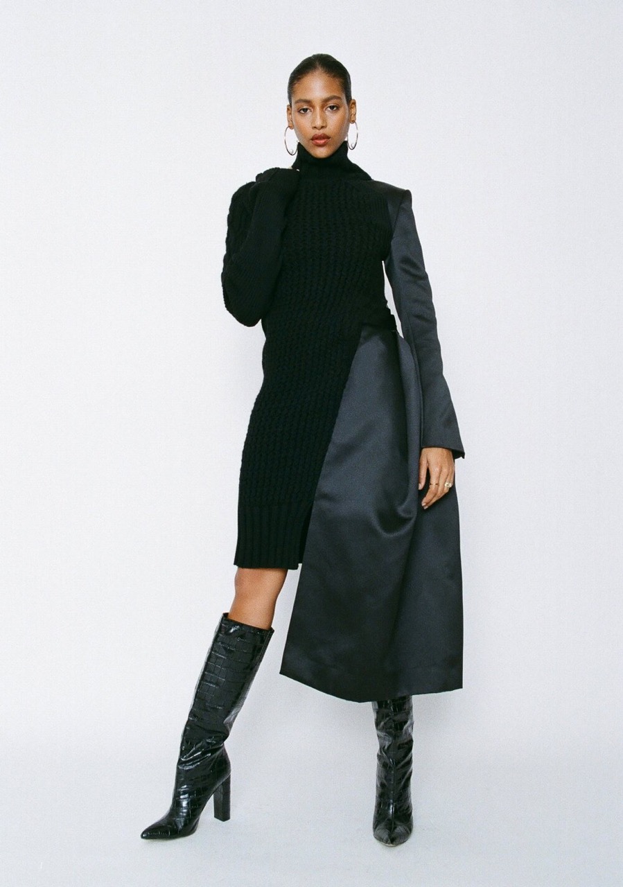 A woman in a black knit dress with a black satin panel and knee-high reptile-texture boots, posing in a full shot against a white backdrop.