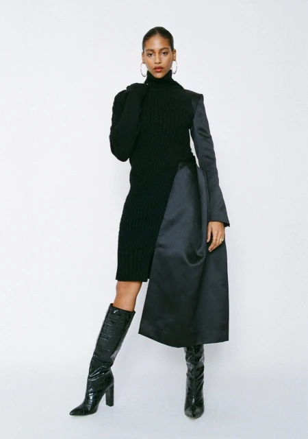 A woman in a black knit dress with a black satin panel and knee-high reptile-texture boots, posing in a full shot against a white backdrop.