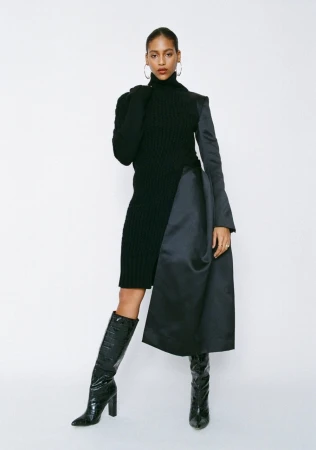 A woman in a black knit dress with a black satin panel and knee-high reptile-texture boots, posing in a full shot against a white backdrop.