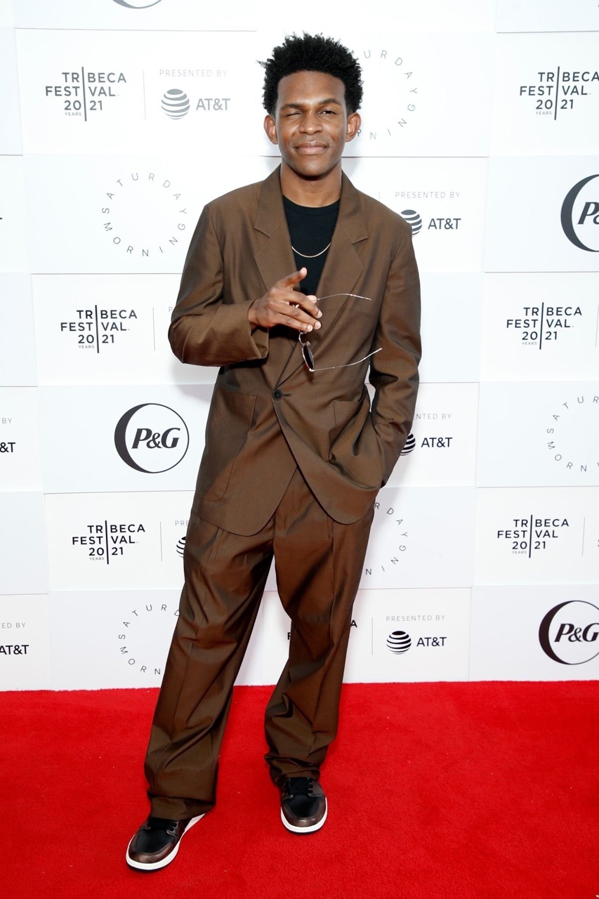 Portrait of a dark-skinned male in a brown suit on a red carpet with event logos in the background; style is modern and fashionable.