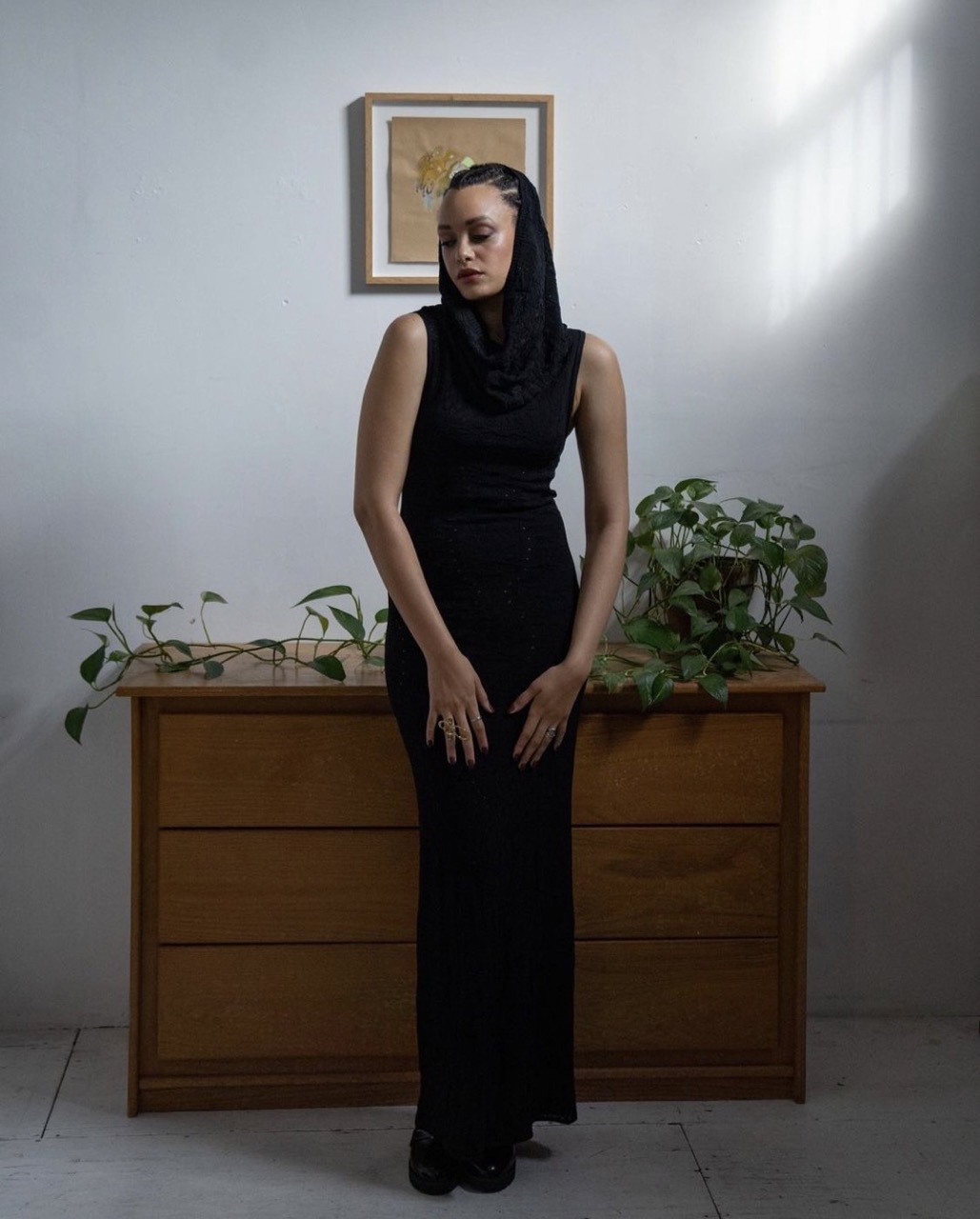 Woman in black dress and head covering poses indoors with natural light, alongside a wooden dresser, plants, and framed artwork.