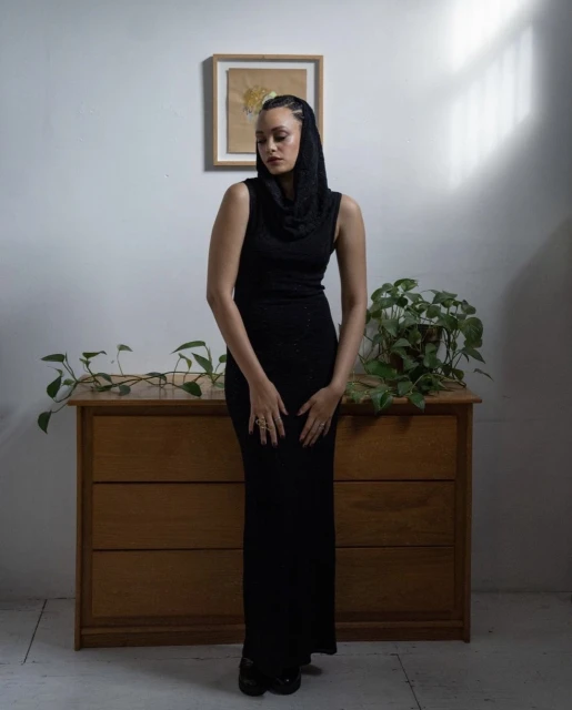 Woman in black dress and head covering poses indoors with natural light, alongside a wooden dresser, plants, and framed artwork.