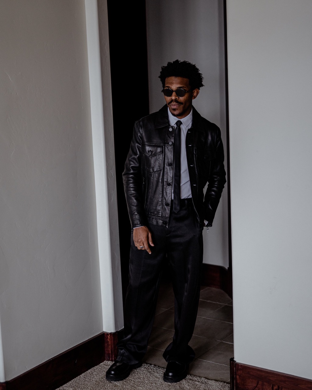 A Black male fashion model strikes a confident pose in a leather jacket and stylish black suit inside a doorway; indoor fashion shot.