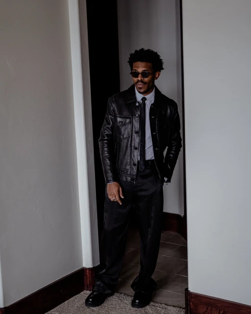 A Black male fashion model strikes a confident pose in a leather jacket and stylish black suit inside a doorway; indoor fashion shot.