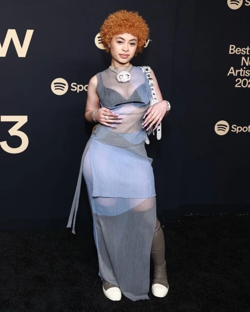 Ice Spice attends the 2023 Spotify Best New Artist Party in a light blue sheer dress and taupe boots, posing in front of a branded backdrop.