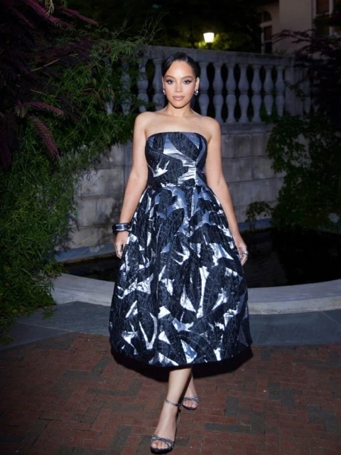 A woman in a strapless, black and silver floral dress stands on a brick walkway in front of greenery and a stone wall.