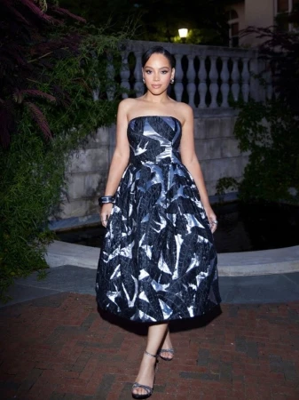 A woman in a strapless, black and silver floral dress stands on a brick walkway in front of greenery and a stone wall.
