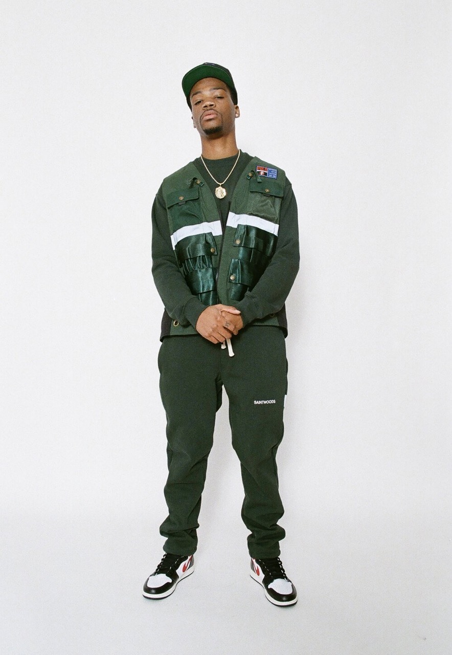 Full-length studio shot of an adult male in an olive green cap, vest, and pants, posing against a white backdrop with hands clasped.