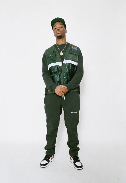 Full-length studio shot of an adult male in an olive green cap, vest, and pants, posing against a white backdrop with hands clasped.