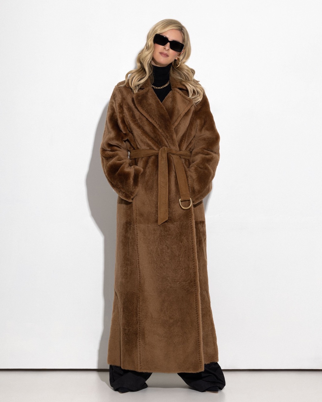Model wearing dark sunglasses and a long brown belted coat against a plain white studio background.