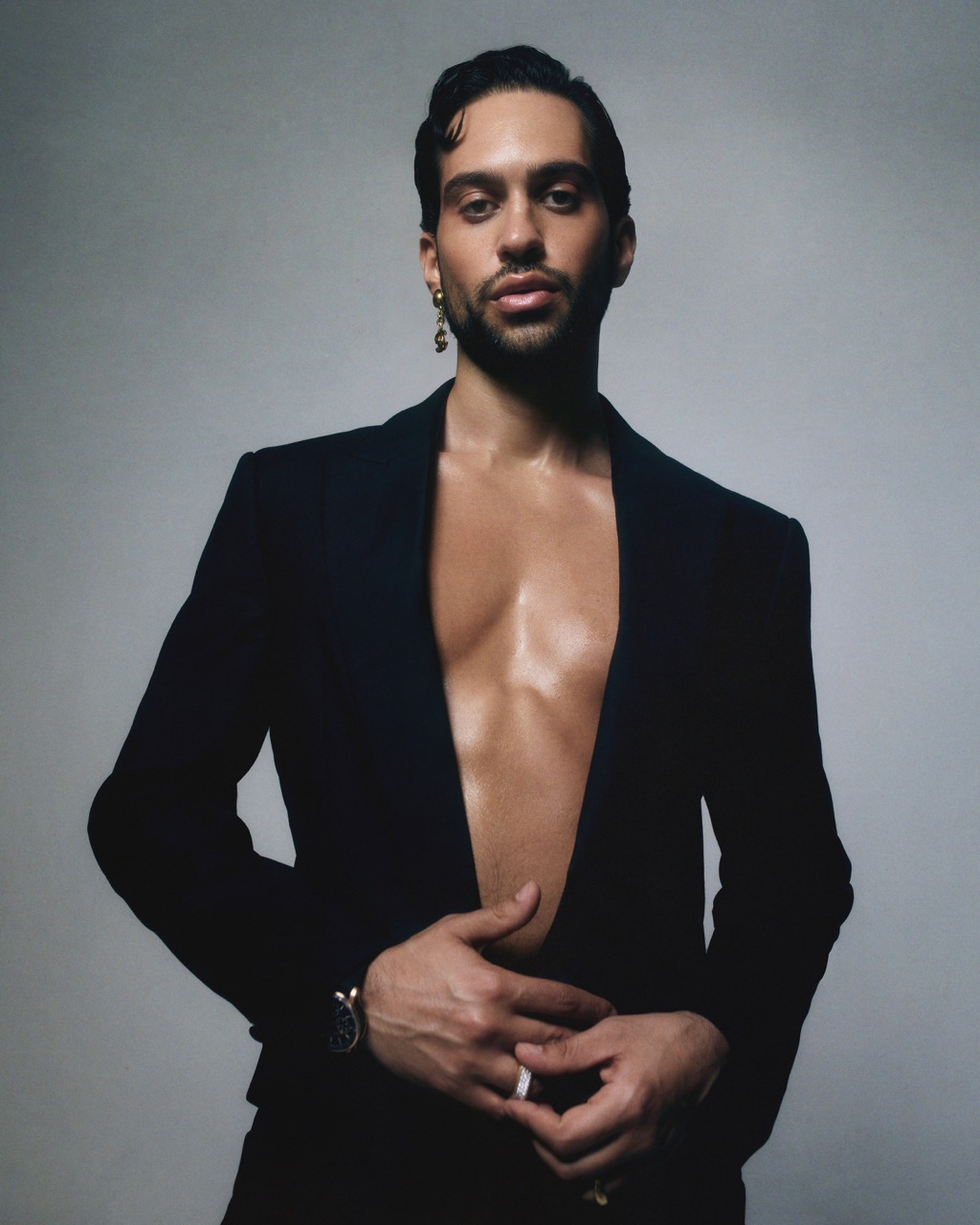 Portrait of a handsome adult male with dark hair and an open black suit jacket, posing against a neutral grey studio background.