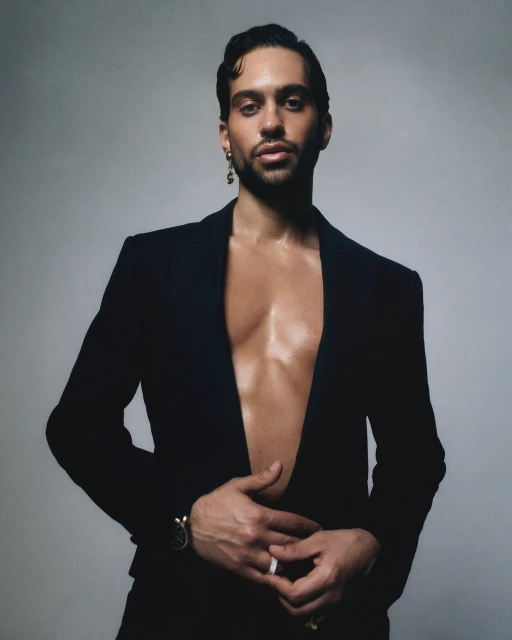 Portrait of a handsome adult male with dark hair and an open black suit jacket, posing against a neutral grey studio background.