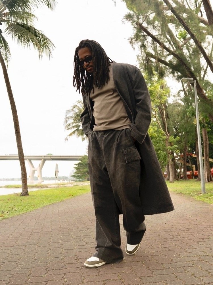Man with dreadlocks in gray coat and trousers walking on brick path along waterfront with bridge and palm trees in background.