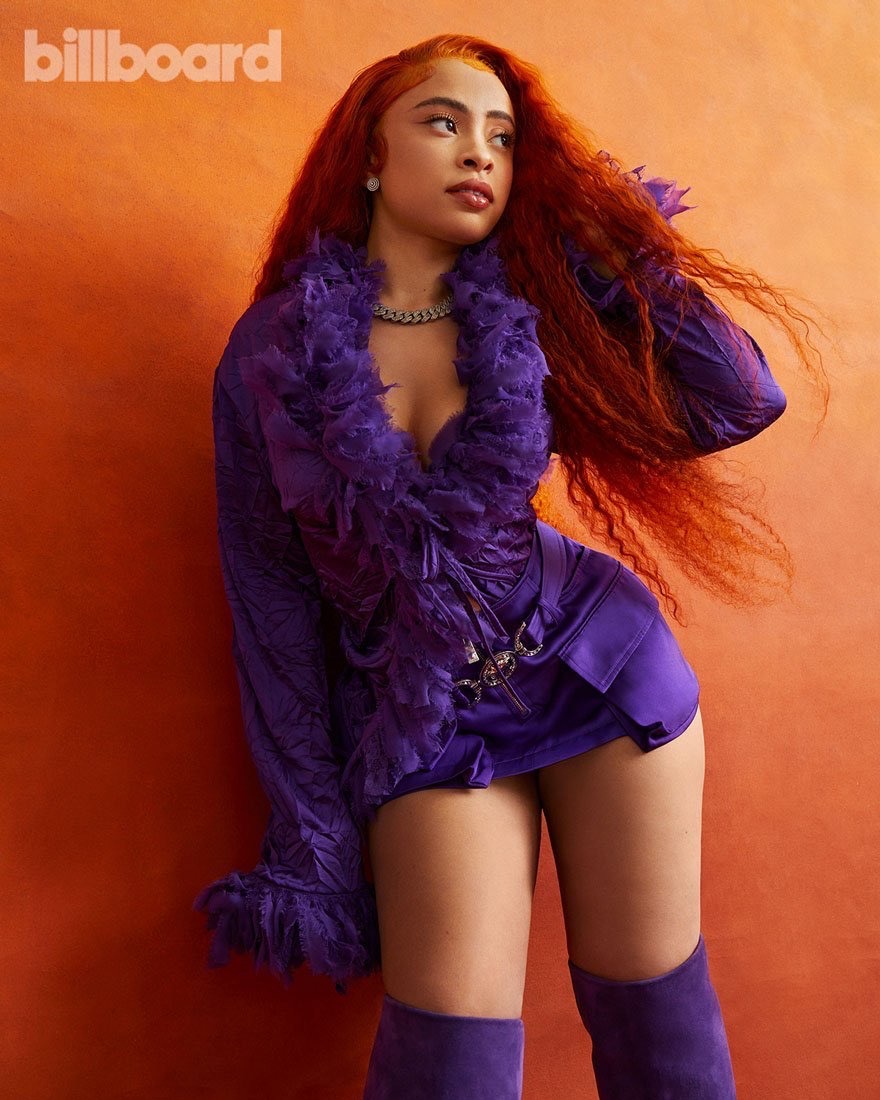 Glamorous woman in a purple outfit poses against an orange backdrop in a fashion-style photograph for Billboard magazine cover.