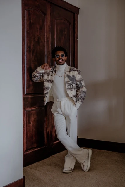 Young man in glasses and stylish printed jacket poses by a dark wood door wearing all-white outfit and sneakers in an indoor setting.