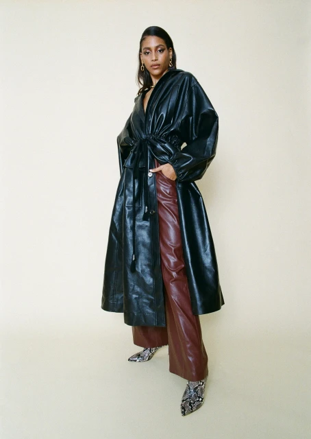 Elegant woman posing in a black leather coat and brown pants in a studio, radiating sophisticated fashion style with natural light.