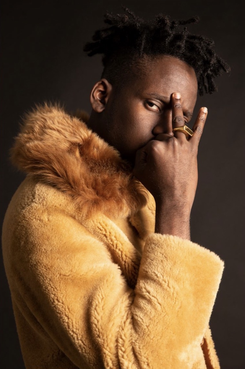 Portrait of a black male in a fur-collared coat against a dark backdrop, striking a hand pose, with focus on fashion and expression.