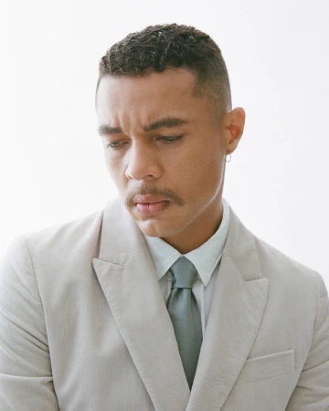 Close-up portrait of a man in a beige suit jacket, white shirt, and tie against a plain white background.