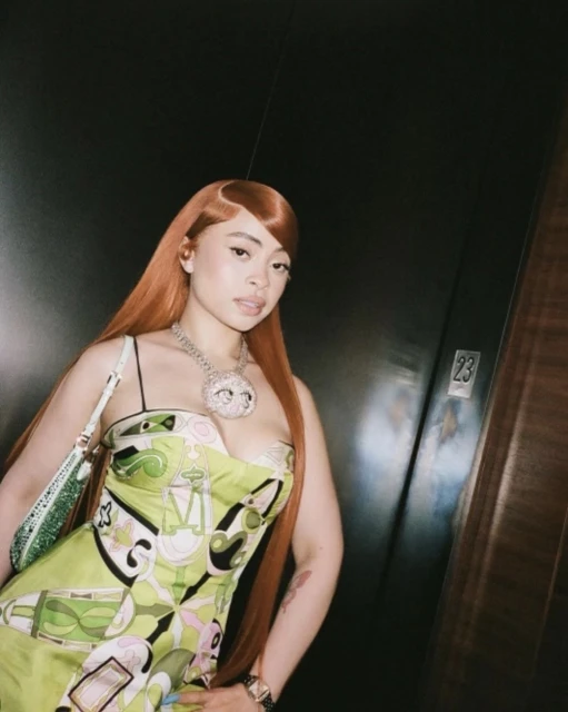 A woman with auburn hair, wearing a floral dress, poses against a dark backdrop with a partially visible door, showcasing fashion and style.