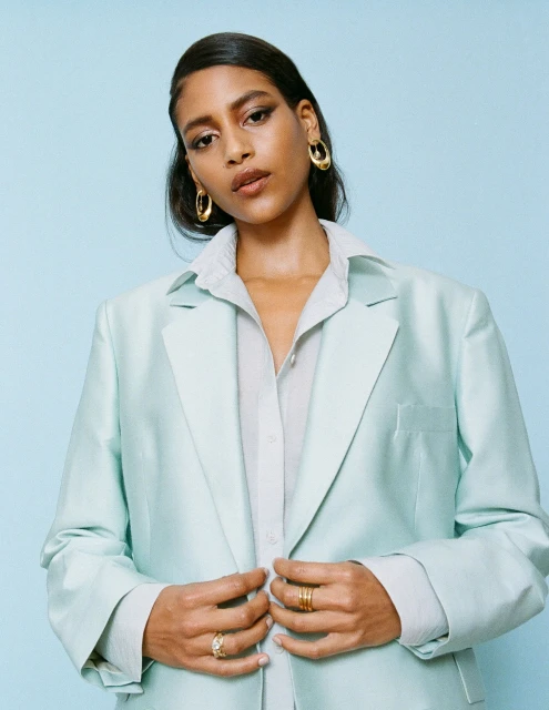 Portrait of a confident woman in a light blue suit and button-down shirt, accessorized with gold hoop earrings and rings, against a blue backdrop.