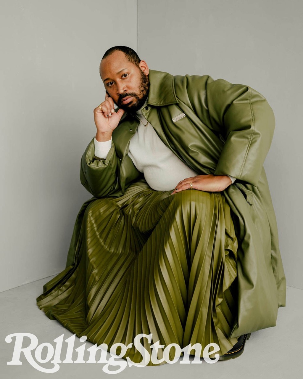 Adult male poses in an olive-green leather coat and matching pleated skirt in a studio setting with a Rolling Stone logo.