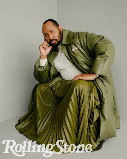 Adult male poses in an olive-green leather coat and matching pleated skirt in a studio setting with a Rolling Stone logo.