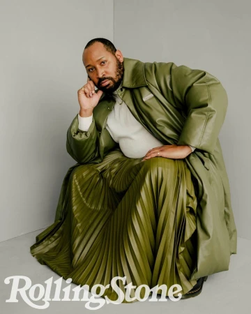 Adult male poses in an olive-green leather coat and matching pleated skirt in a studio setting with a Rolling Stone logo.