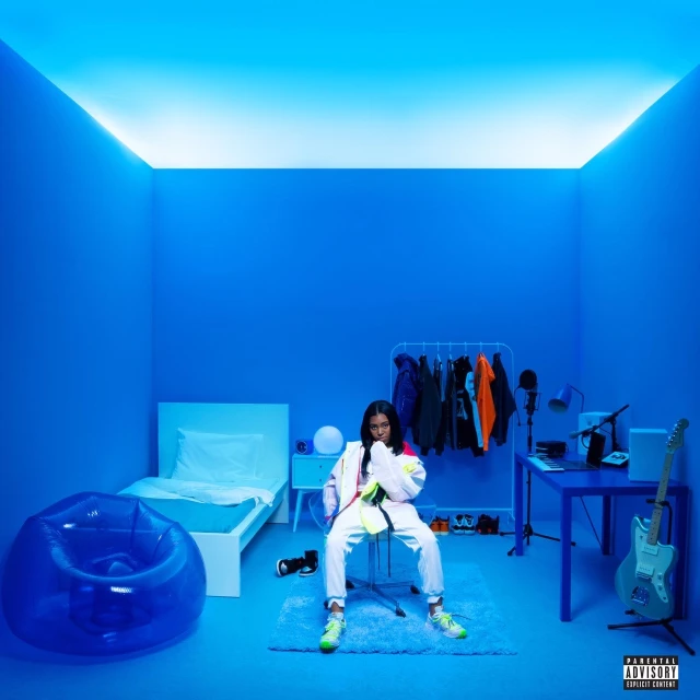 Teenage girl sitting on a chair in a blue room with recording equipment, a bed, and blue inflatable chair for youthful lifestyle content.