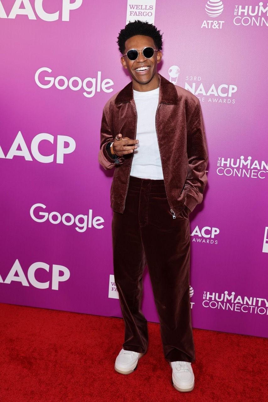 A smiling adult male on a red carpet wearing a brown velvet suit, white shirt, sunglasses, and white sneakers attends the NAACP awards.