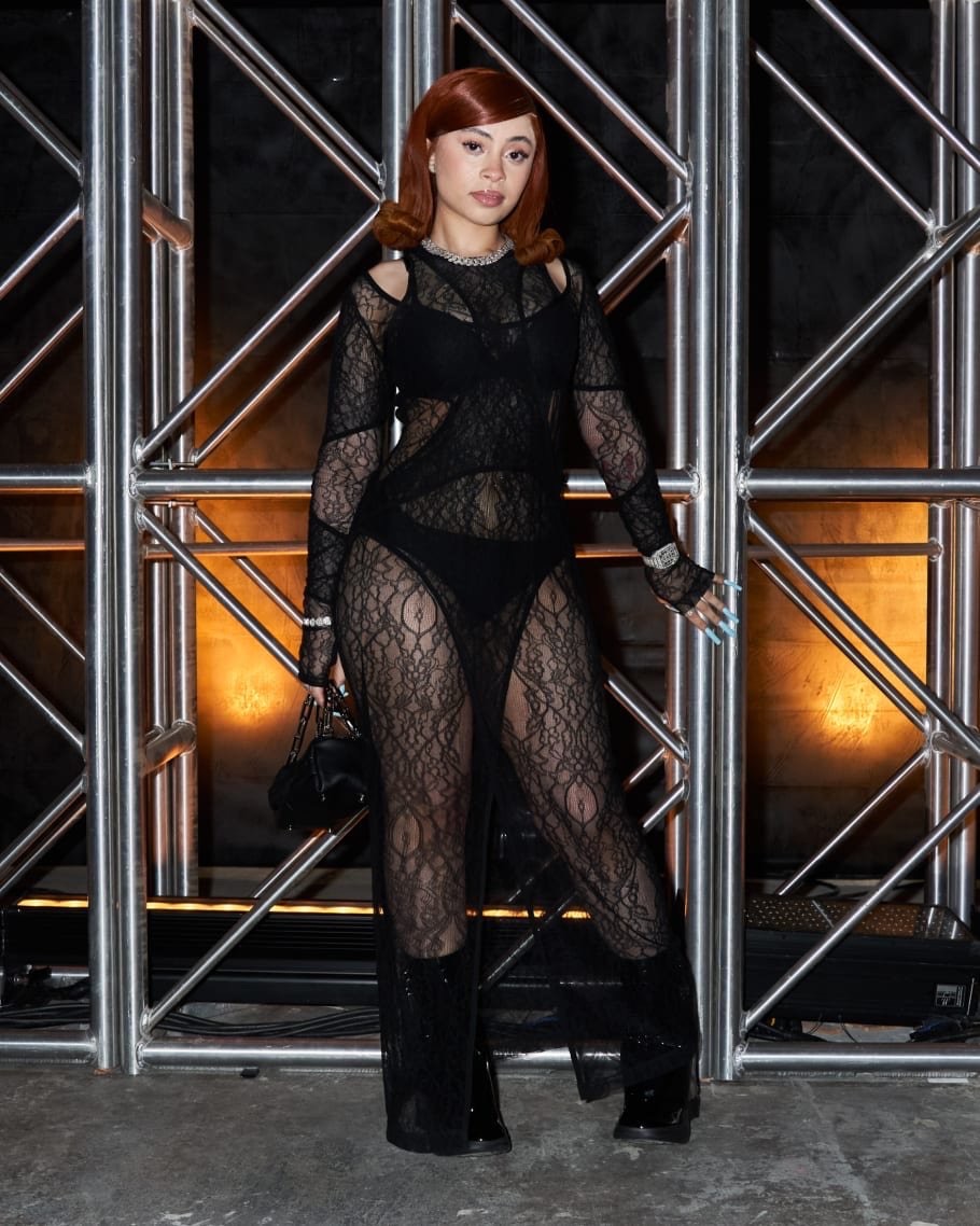 Ice Spice poses in a black lace catsuit and platform boots against a steel-beamed backdrop, showcasing a bold fashion-forward statement.